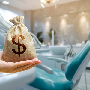 A hand holding a money bag in front of a teal dental chair